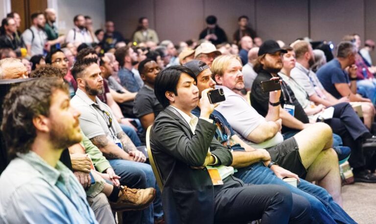 A large audience sits in rows at a conference, with some attendees holding up phones to record the event.