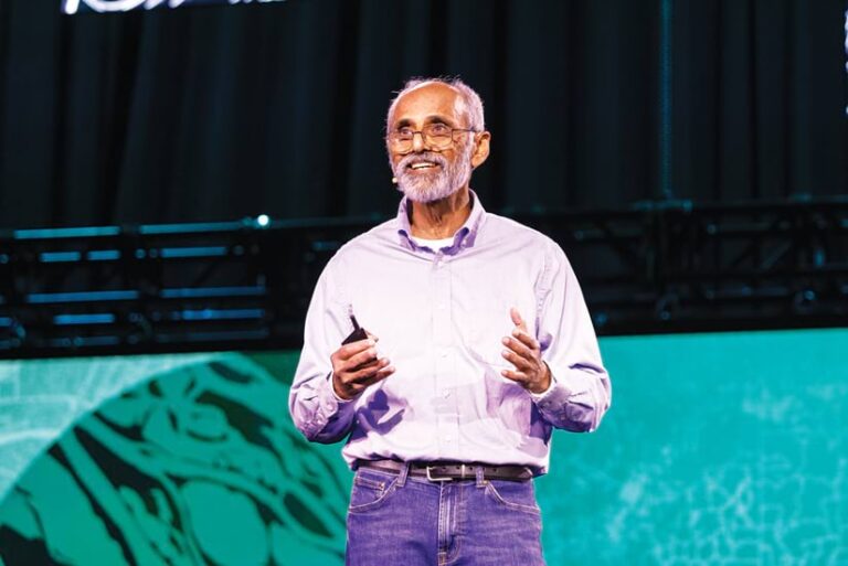 A man with a gray beard and glasses stands on stage, smiling and gesturing with a clicker in hand. He wears a light purple button-down shirt and jeans. Behind him is a large screen with a green and black design.
