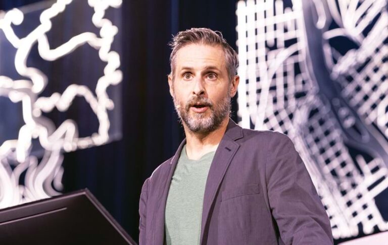 A middle-aged man with short graying hair and a beard speaks on stage, wearing a dark blazer and green t-shirt. Abstract white lights glow in the background.
