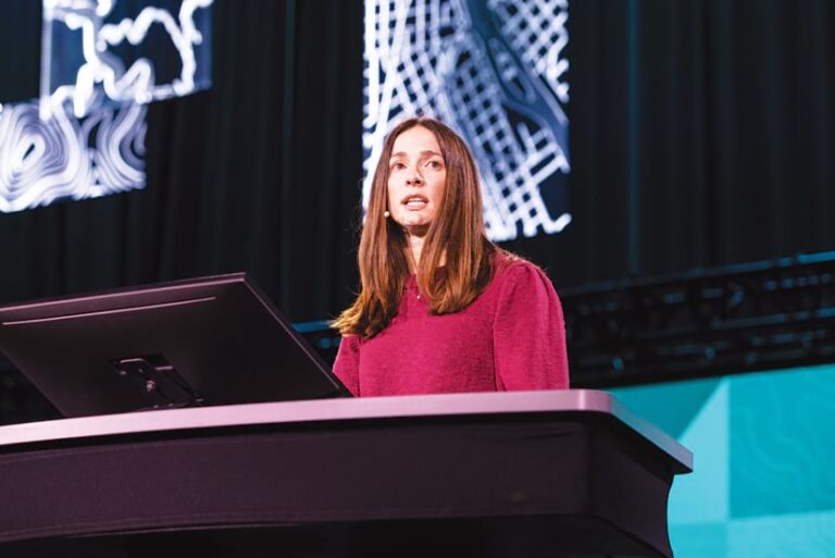 A woman in a magenta sweater speaks at a podium on stage, with illuminated map graphics behind her.