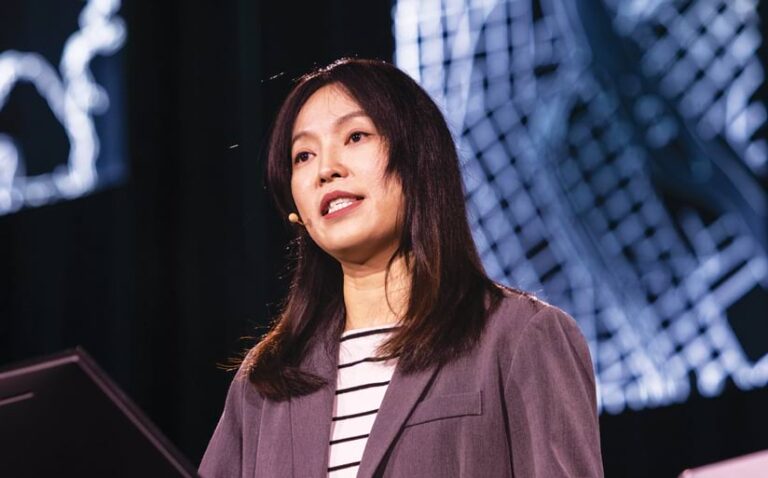 A woman with dark hair, wearing a gray blazer and a small microphone, speaks on stage in front of a large abstract projection.