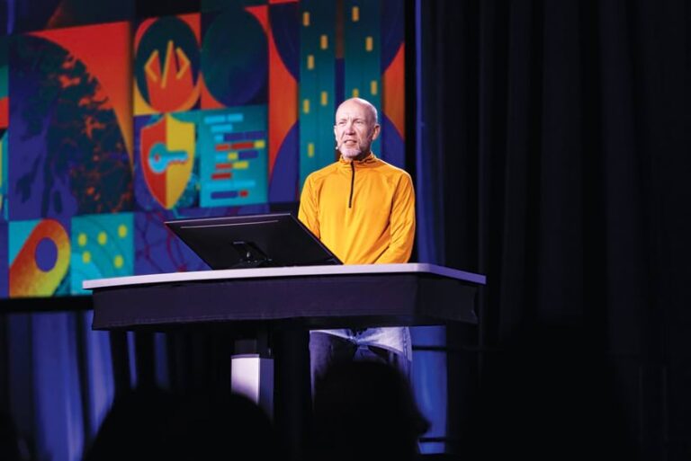 A bald man in a yellow pullover speaks at a lectern on stage. Behind him, a large screen shows a colorful abstract graphic with technology icons.