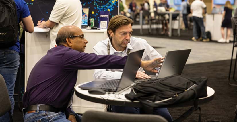 Two men sit at a small table with laptops in a busy conference hall. The man in a purple shirt and glasses points to the other man's laptop screen.