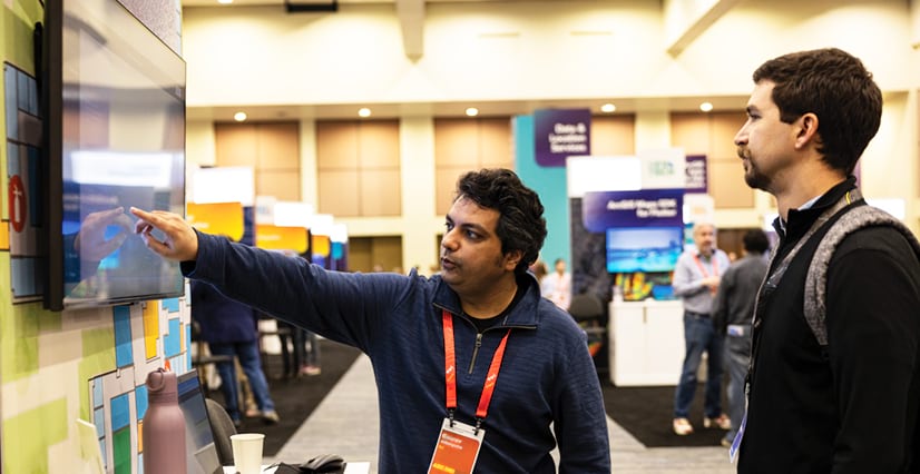 A man in a blue pullover points at a large touchscreen monitor while explaining something to another man at a conference.