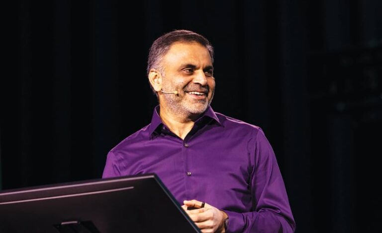 A smiling man in a purple button-down shirt stands at a lectern on stage, wearing a small over-the-ear microphone.