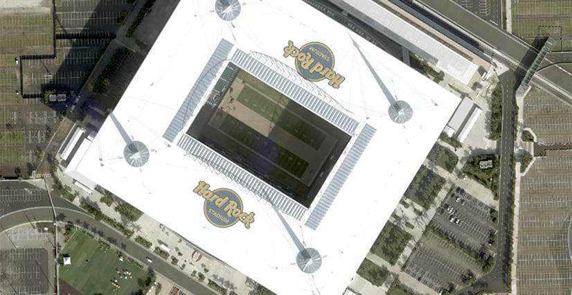 An aerial, top-down view of Hard Rock Stadium with a large white roof, a rectangular opening over the green field, and the "Hard Rock Stadium" logo. Extensive parking lots surround the stadium, and umbrellas dot the grassy areas outside the stadium entrance.