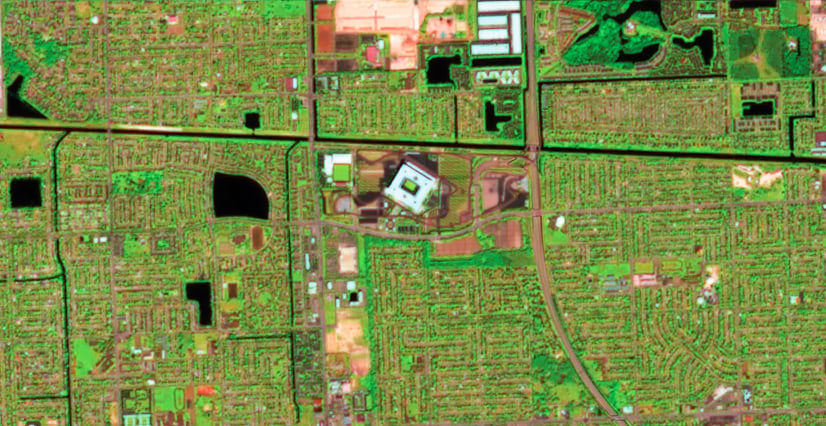 A false-color satellite image of a suburban grid with a large white stadium at the center. Streets and houses form a pattern around the stadium; vegetation appears bright green, and water areas are black.