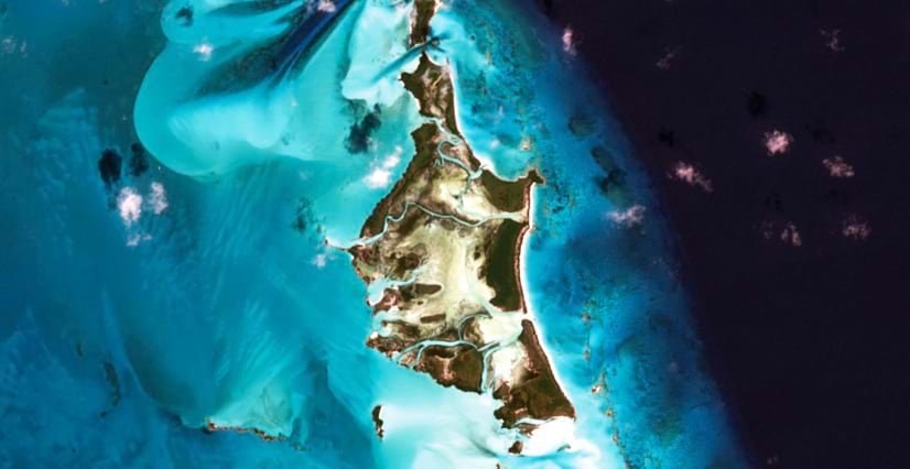 A satellite image of a tropical island with green and brown land surrounded by swirling turquoise water and deep blue ocean.