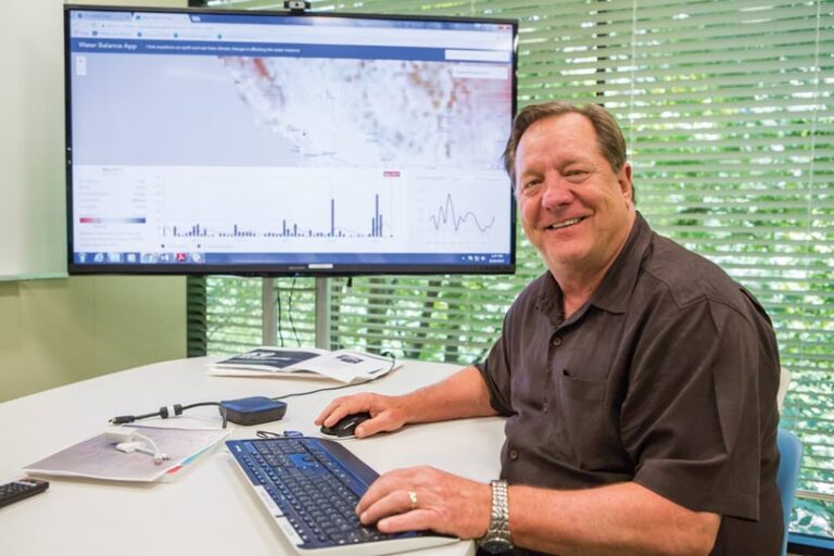 Man sitting at a computer, smiling at the viewer, with mapping software displayed on the monitor.