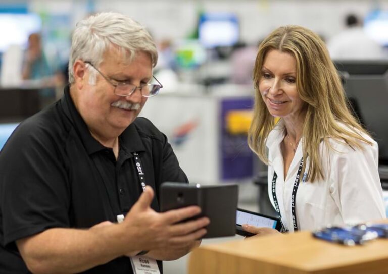 Man with a mustache holds up a tablet in front of a woman, showing her something on the screen.