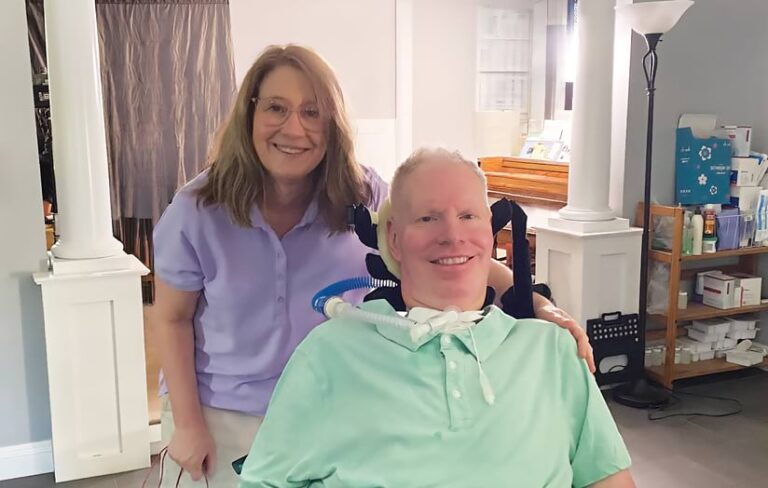 A smiling man with a tracheostomy tube sits in a wheelchair. A smiling woman stands behind him with her hand on his shoulder.