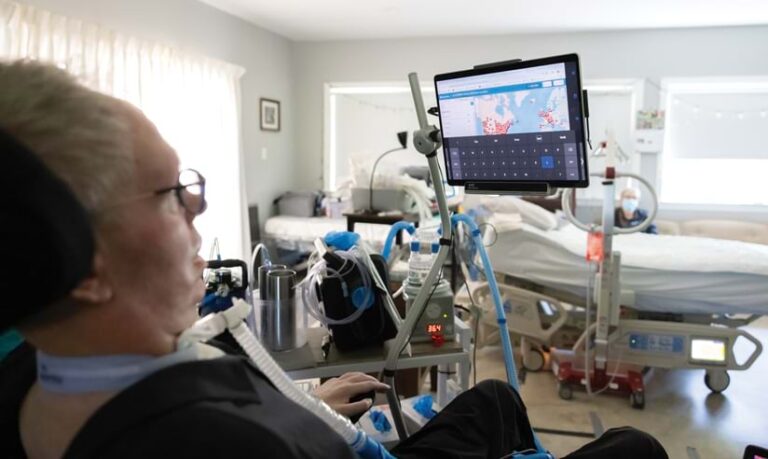A person with a tracheostomy tube sits in a specialized chair, using a tablet mounted on an assistive device that displays a world map.