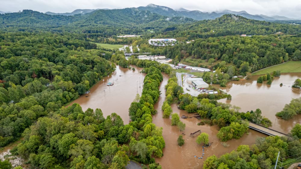 Aerial view of brown floodwater covering roads and buildings in a wooded river valley after Hurricane Helene.