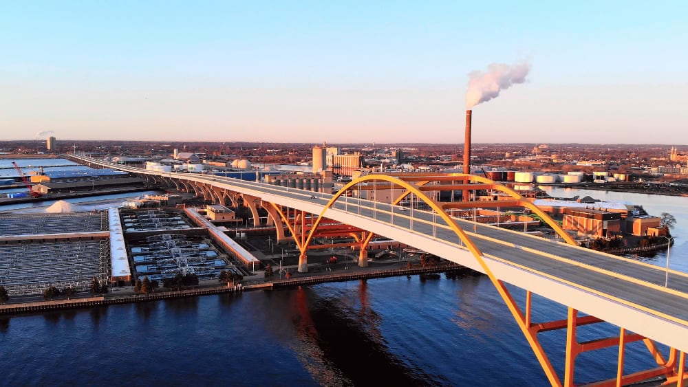 Aerial view of a wastewater treatment plant beneath an arched bridge with a smokestack and city skyline.