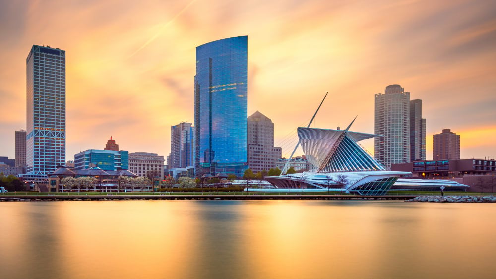 Milwaukee skyline at golden hour reflected in calm lakefront water, with the Art Museum in view.