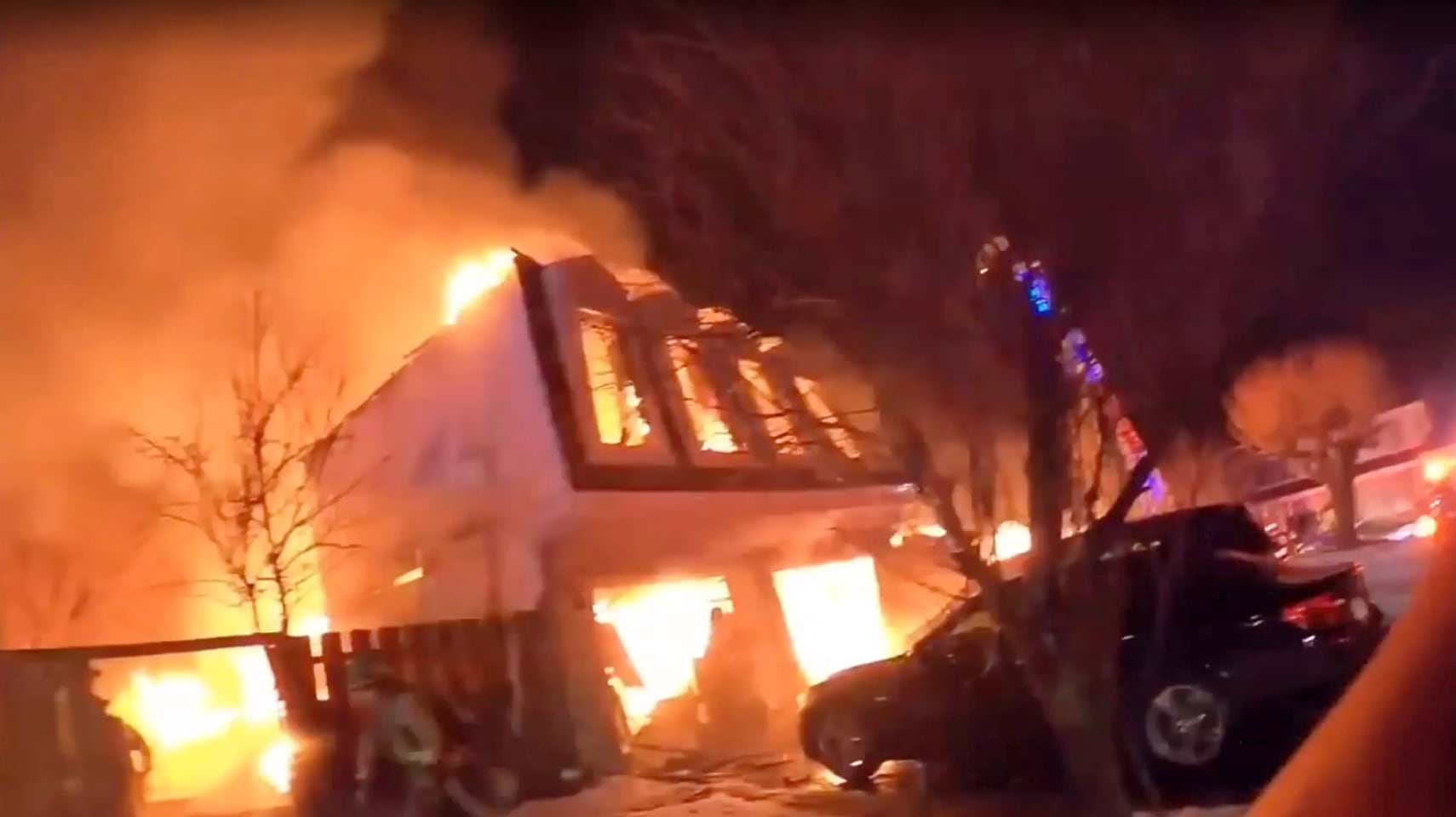 House fully engulfed in flames at night, firefighters and a damaged vehicle in the foreground.