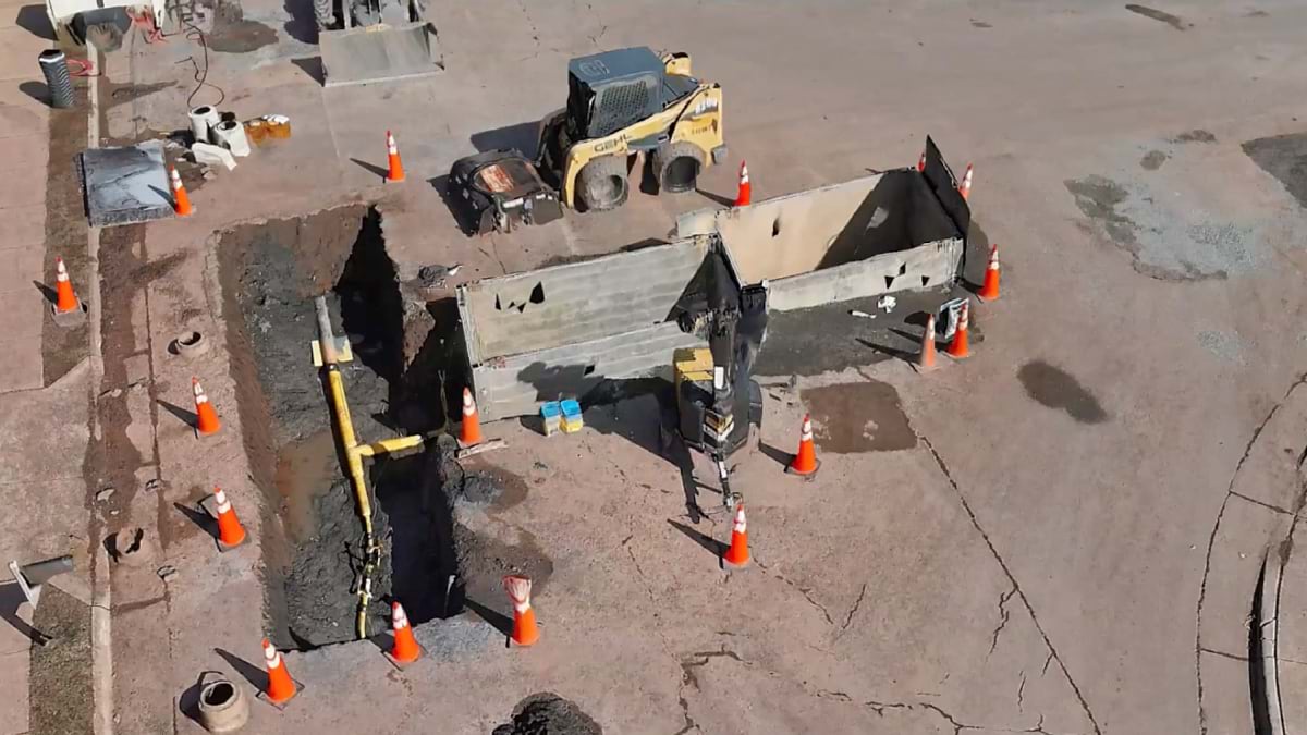 Aerial view of an excavated trench with a backhoe and safety cones marking a gas line dig site.