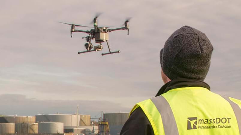 MassDOT Aeronautics Division pilot in yellow safety vest watches a multirotor drone hover over an industrial facility against a cloudy sky.