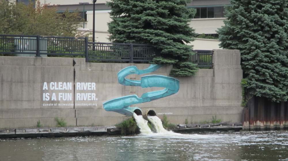 Mural reading "A clean river is a fun river" above two outflow pipes discharging into an urban river.