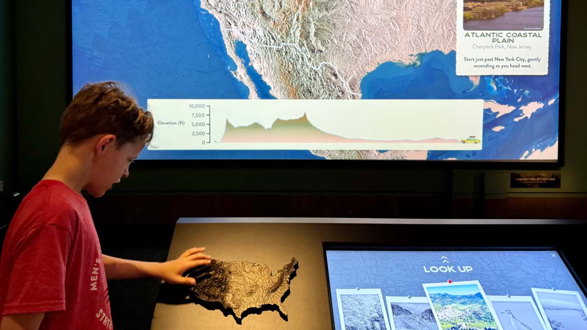 Boy touching a raised relief model of the U.S. Behind him, a screen shows an elevation profile from New Jersey heading west across the country.