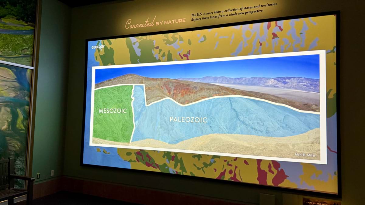 Wall screen titled Geology showing a desert mountain photo overlaid with colored regions labeled Mesozoic and Paleozoic, marking rock ages.