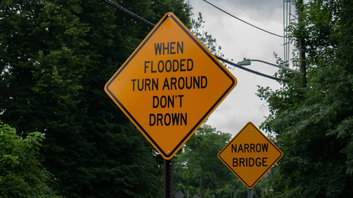 A yellow diamond road sign reading "When Flooded Turn Around Don't Drown" next to a narrow bridge sign.
