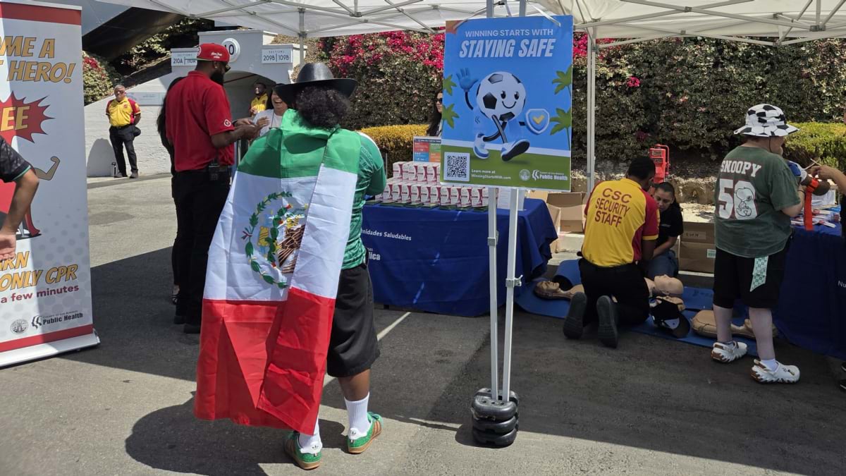 Fan in a Mexico flag walks past a CPR training demo and a Winning Starts With Staying Safe sign at a public health booth.