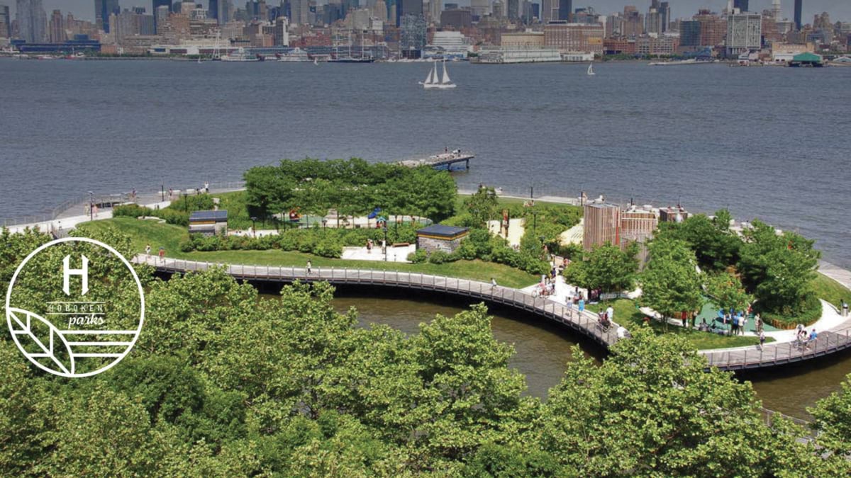 Aerial view of a landscaped Hoboken waterfront park jutting into the Hudson River with green lawns, walking paths, and the Manhattan skyline behind.