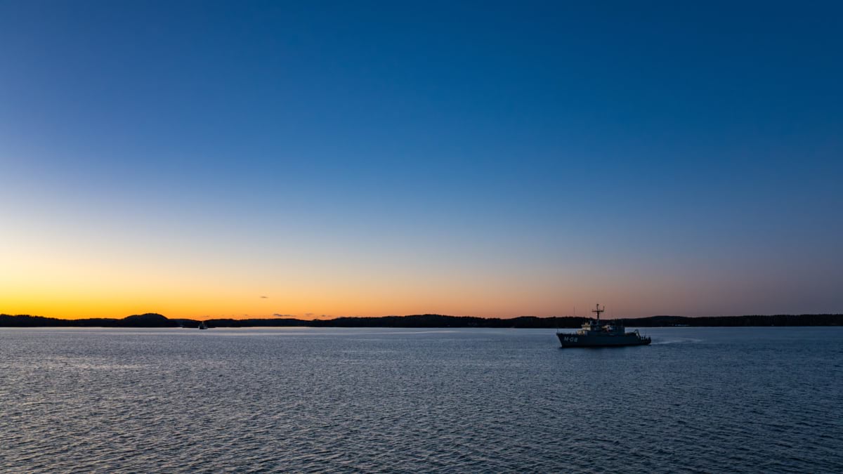 A Latvian minehunter ship sails across calm water at sunset during a NATO naval exercise.