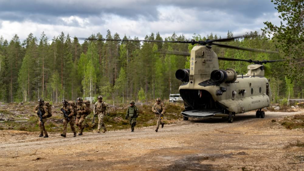 Soldiers from multiple countries climb out of a military helicopter during a NATO exercise in a forest.