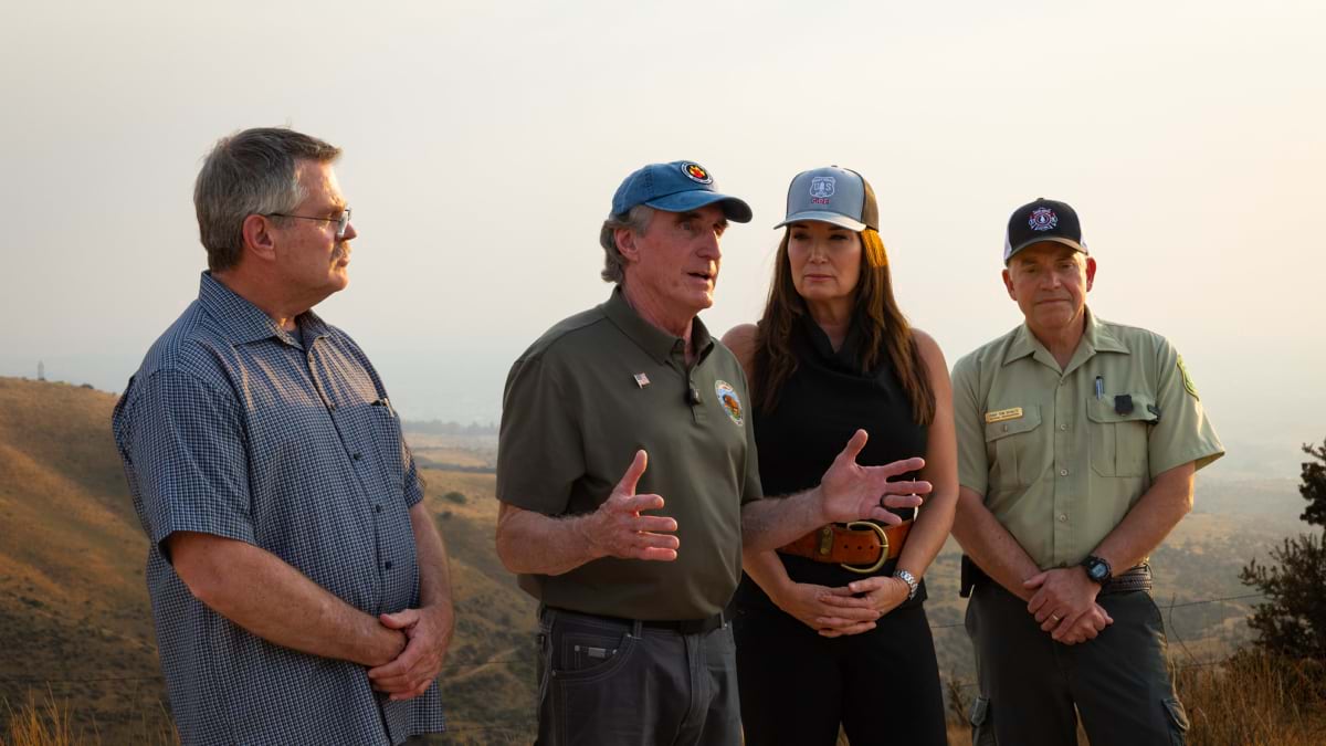 Four officials in fire-agency caps and gear stand on a hillside overlooking smoky terrain, in conversation.