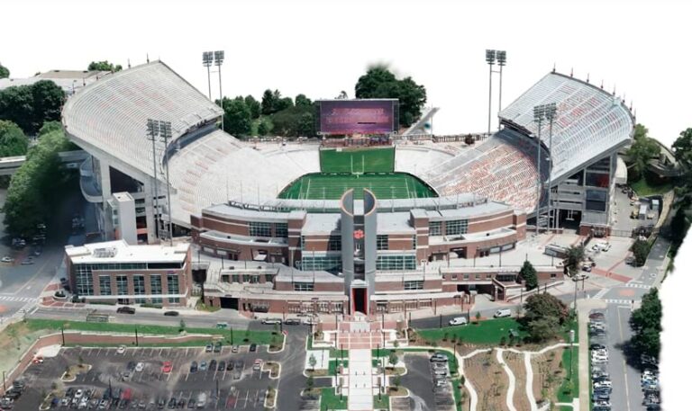 A realistic, Gaussian splat-made aerial view of a large, horseshoe-shaped football stadium with a modern brick exterior and a central entrance tower.