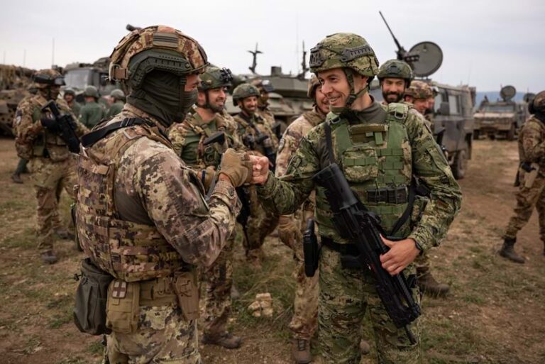 Two soldiers in full combat gear smile and fist-bump in a field, with other soldiers and military vehicles in the background.
