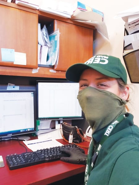 A photo of Terese Flores sitting at a computer wearing a green mask that matched her green CERT hat and shirt