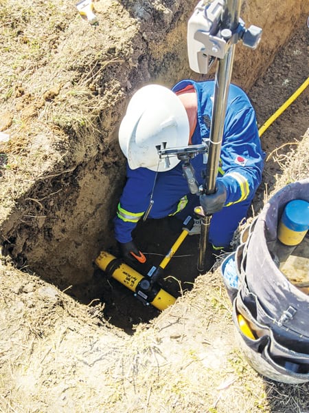 A photo of a fieldworker in a blue suit and a hardhat taking a bar code measurement of an underground asset that has yet to be buried