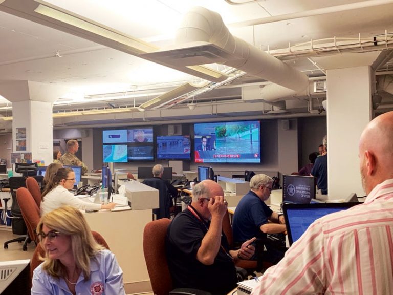 People in the State Emergency Operations Center looking at computers and various electronic displays that show dashboards, news, and maps