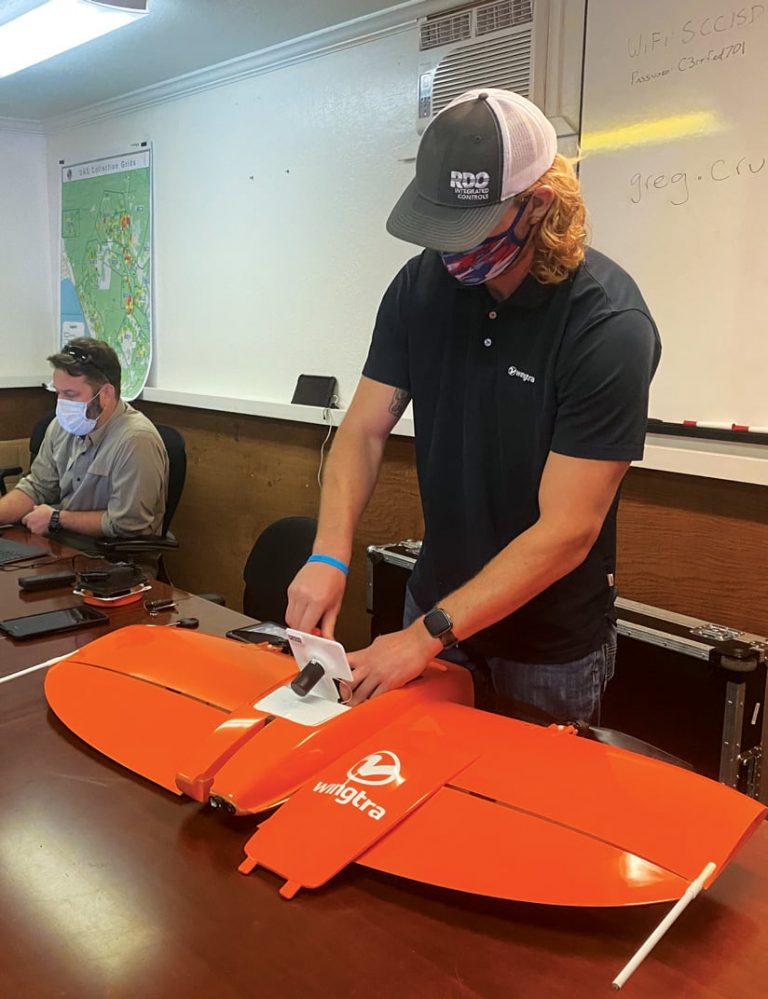 A person with a face mask and baseball cap builds an orange drone in an office