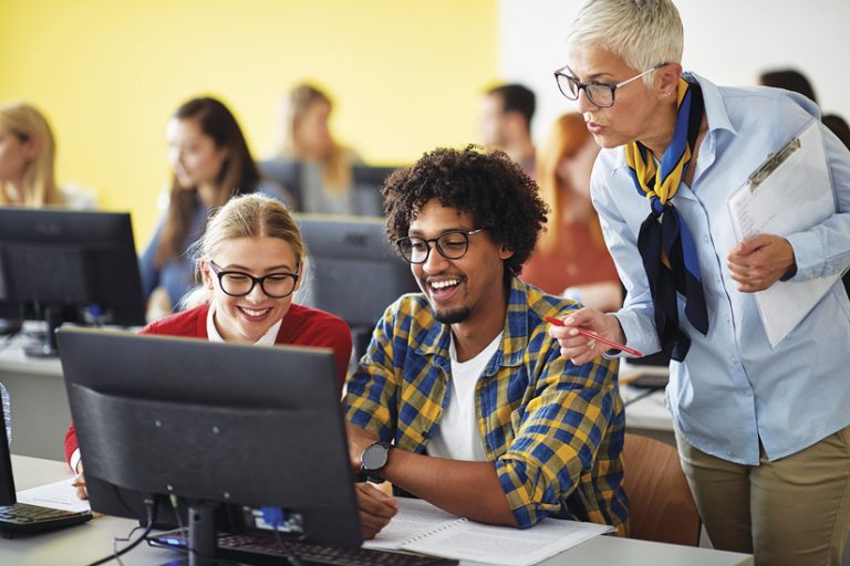 A teacher leaning over the desk of two students who are working at a computer