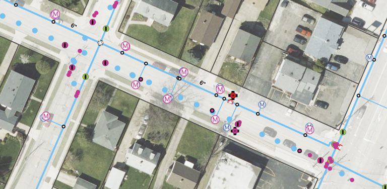 An aerial image of a residential neighborhood with blue lines; blue, pink, and green dots; and pink and purple Ms that indicate various utility assets