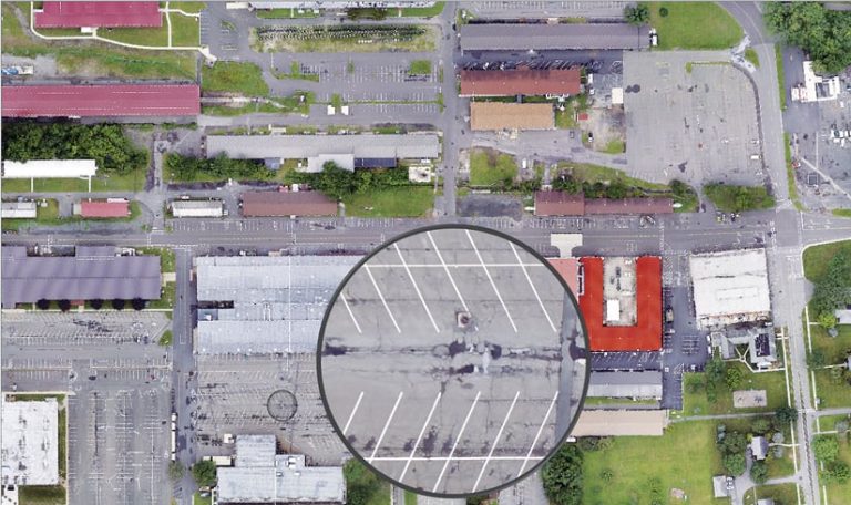 Aerial imagery of a small section of a town, showing some streets, parking lots, and buildings, with one area of the image zoomed in on a parking lot with broken asphalt