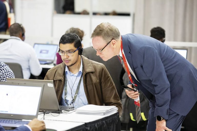 Two men at a laptop—with one using it and one hovering over, helping—in a room where several other people are using laptops