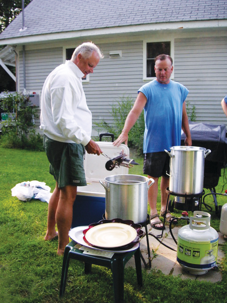 Two men standing near two big, silver pots, with one of them holding a lobster in some tongs