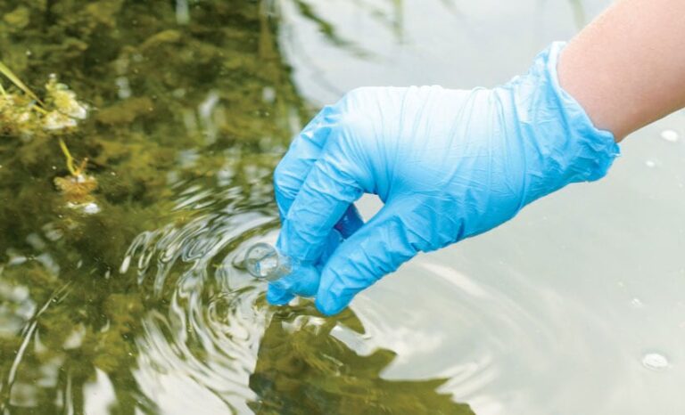 A gloved hand using a beaker to take a water sample from a river