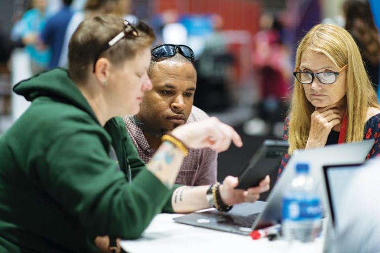 Three people huddled around a laptop and a mobile device, with two of them listening to what the other one is saying