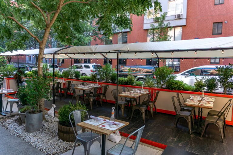 Outdoor tables at a restaurant extending into the street, surrounded by plants and covered by a nicely lit tent