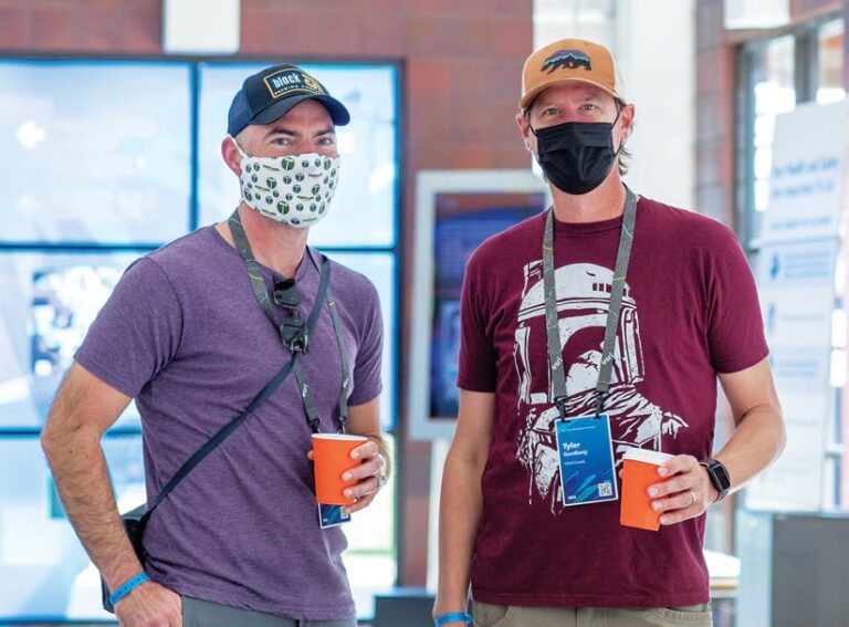 Two DevSummit attendees wearing baseball caps, T-shirts, and masks holding coffees while smiling at the camera