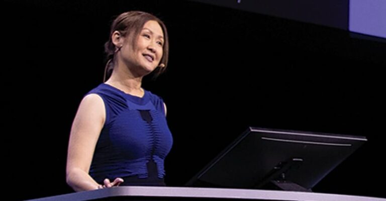 Jianxia Song in a blue dress speaking at a podium