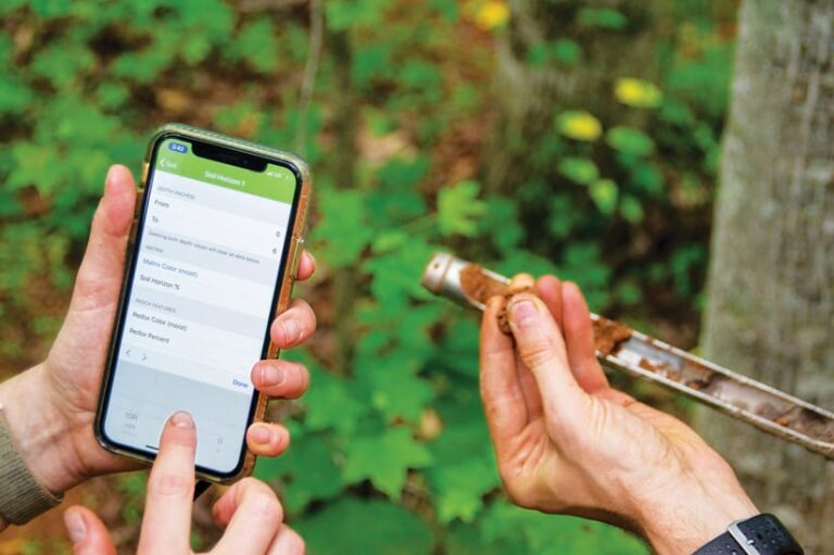 Someone’s hands filling out a soils report on a smartphone while someone else holds dirt up in one of their hands