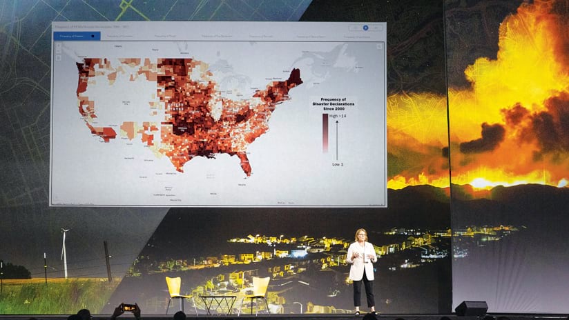 A woman onstage speaking in front of a red-colored map of the United States and a big photo of a wildfire