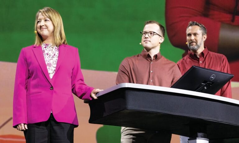 A woman in a pink blazer standing onstage next to a table and two men in red shirts standing behind the table